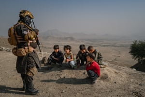 A young boy sat on his haunches looks up at an armed police officer in combat fatigues while four others play in the dirt on a ridge overlooking an arid mountain valley