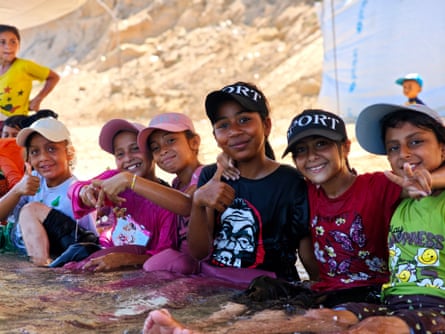 A group of children sitting at the water’s edge, some of them giving the thumbs up and other the peace sign