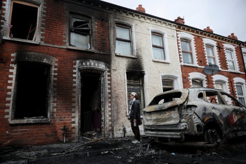 A woman stands on the pavement outside her fire-blackened house. A burned-out car is also in the picture