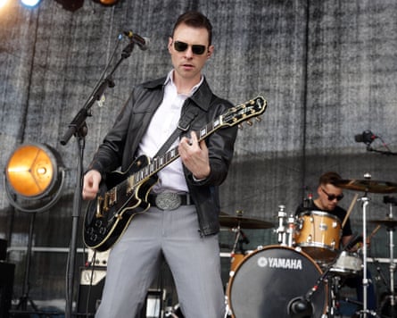 Man in sunglasses plays electric guitar on stage with drummer behind
