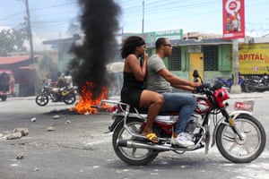 A man on a motorcycle and a woman riding pillion and covering her nose and mouth, ride past a plume of black smoke