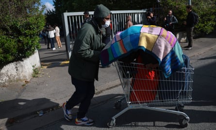 A man pushes his belongings in a supermarket trolley to take a bus; he wears a dark anorak and hood and his things are covered by a colourful knitted blanket.