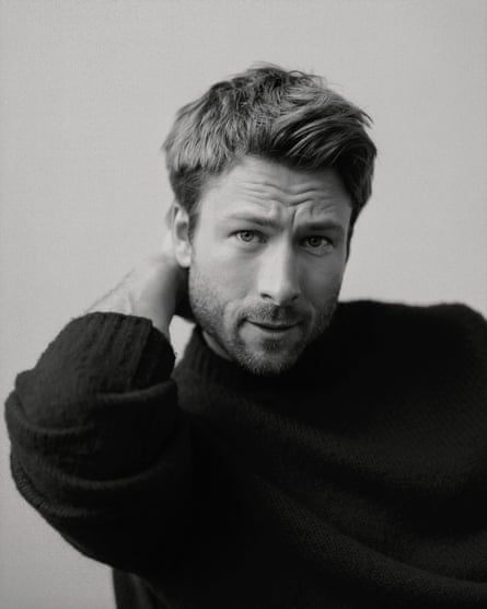 Portrait of Glen Powell looking at the camera with this right arm bent and his right hand on the back of his neck wearing a black jumper