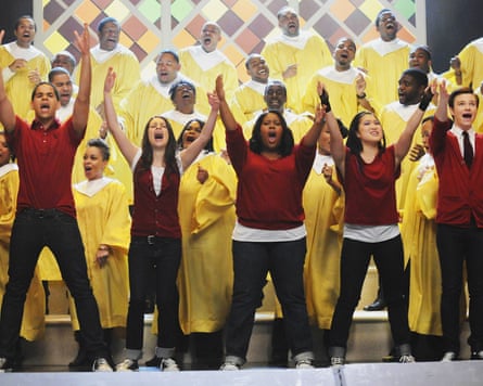Row of high schoolers stand with arms in the air as they sing on stage in front of choir with yellow robes