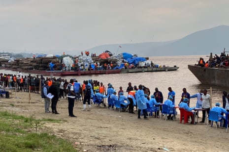 A long line of people queue to be tested by health workers sitting at tables next to a river