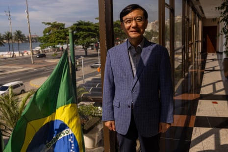 A man in a suit poses in front of a Brazilian flag near a beach