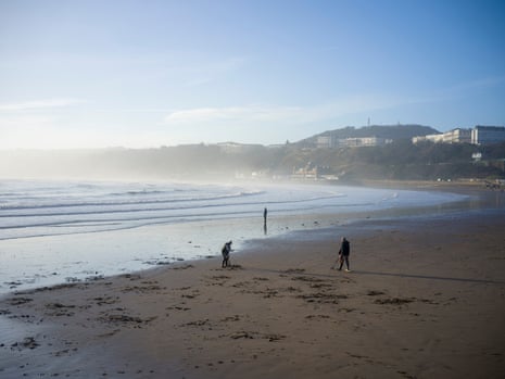 A view of two metal detectorists on a sandy beach in a wide bay with another person in the distance staring out to sea