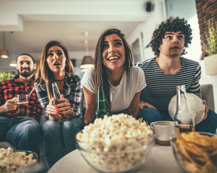 Group of sports fans sitting on a sofa surrounded by snacks and drinks.