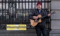 Martin Leahy strumming a guitar next to a yellow sign that reads: #HousingCrisis.