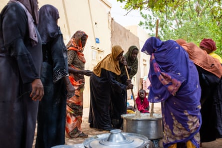 Women wearing hijabs serve meals from large vats to a line of people waiting outside on a street