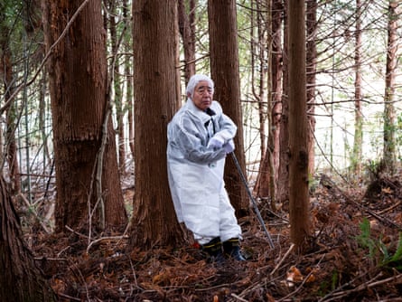 A man dressed in a white protective suit and hat leans against a tree in a forest landscape