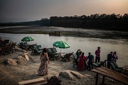 A few people sit at tables and chairs next to a body of water in low light. Others stand around. Trees can be seen on the other side of the water