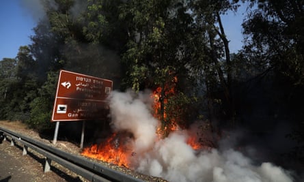 Flames and billowing smoke are seen alongside a hillside road with trees behind; the smoke almost covers a large road sign. The sky is blue in the background, in sharp contrast to the bright orange flames and dark green of the trees.