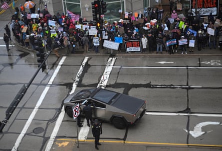 Police officers stand next to a Cybertruck parked in the middle of a road as a crowd of protesters with signs stand on the nearby sidewalk
