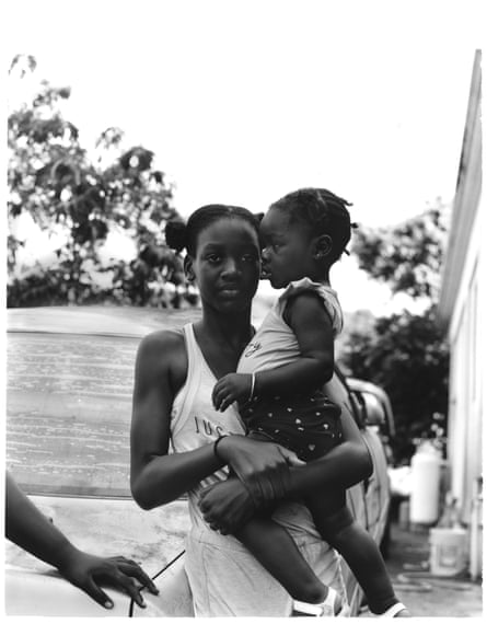 A young person holds a younger child while leaning against the rear wing of a car