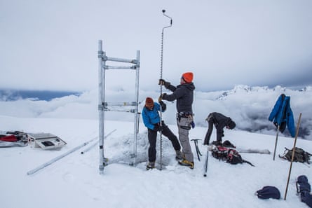 Two people on an icy peak with a cloud behind them hold a long metal piece of equipment. A third person bends over behind them. Other pieces of equipment and clothing can be seen around them
