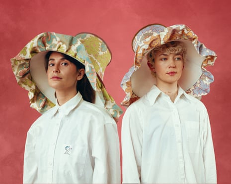 Charly Clive and Ellen Robertson in white shirts wear large decorative hats with ruffled floral brims