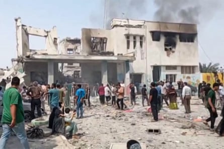 People stand in front of a partially demolished building with smoke billowing from one section.