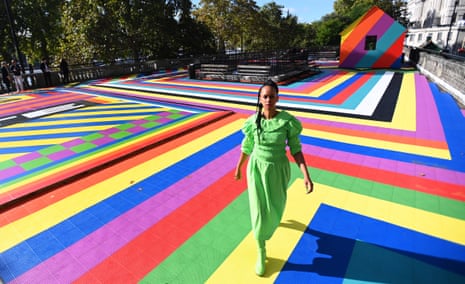 Artist Lakwena stands on a colorful striped rooftop installation with a rainbow structure behind
