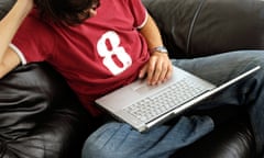 Male wearing red t shirt and jeans sitting on sofa using laptop