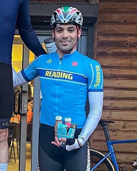 close-up portrait of Mohammad Ganjkhanlou, seen with his arm around another cyclist on a podium; he wears a light blue club shirt, black shorts and a helmet, and is holding two bottles which look like energy drinks and some packets which look like high-energy foodstuffs. A blue racing bike is seen in the background against a wooden-panelled building