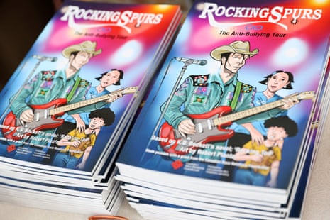 Two piles of soft-bound books with colorful graphic illustration of man wearing cowboy hat playing electric guitar, woman behind him and child.