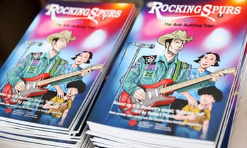 Two piles of soft-bound books with colorful graphic illustration of man wearing cowboy hat playing electric guitar, woman behind him and child.