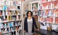 Reena Barai: she is standing in a doorway between shelves of medicines in packaging, and wears a black and white striped dress with black cardigan. She is Asian and has collar-length curly dark hair and glasses; she is smiling and resting one hand on a black desk