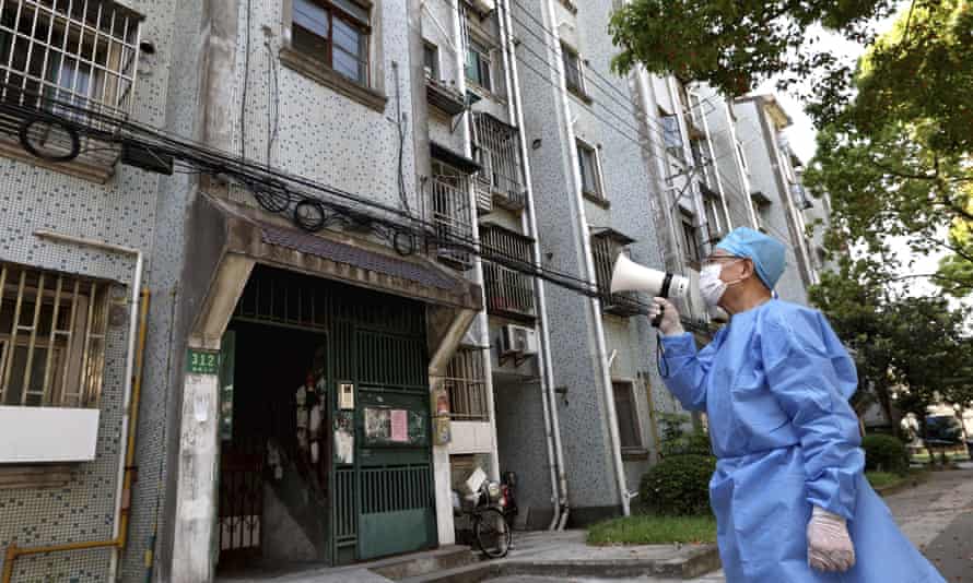 A man in full protective clothing and a mask standing in the street, using a megaphone, in front of a modern block of flats