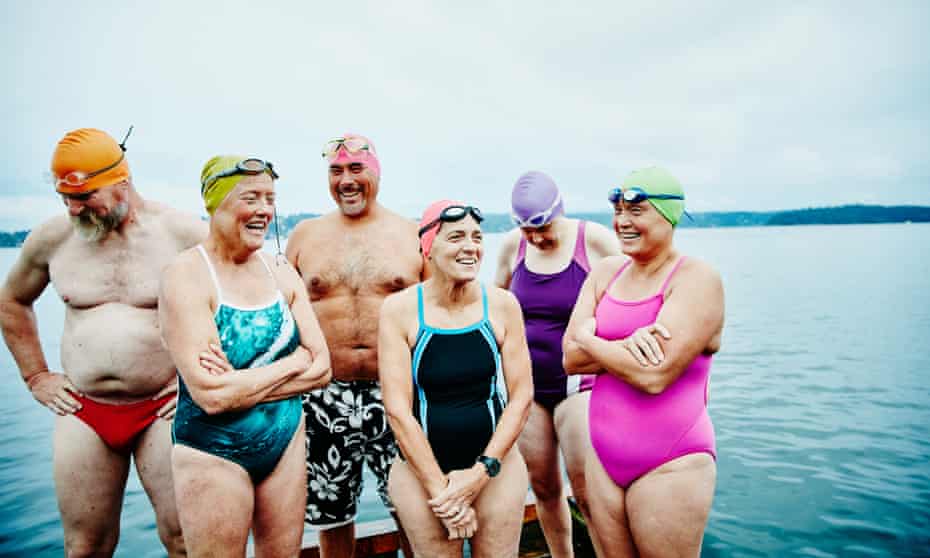 Six older people in swimming costumes, hats & goggles stand on a dock by a lake
