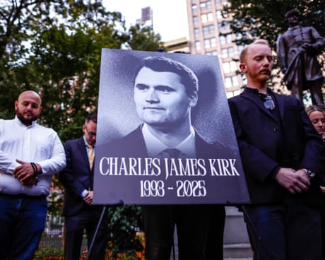 people stand with their heads bowed around a sign with a picture of a man that reads 'Charles James Kirk 1993-2025'