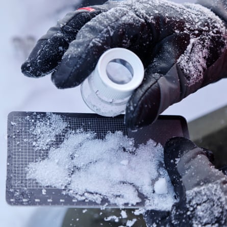A gloved hand holds a magnifier to snow crystals on a card