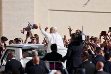 The pope waves to crowds of people from the open top of a car which is surrounded by men in suits. One person in the crowd holds a baby above their head
