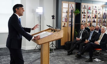Rishi Sunak delivers his speech at the Policy Exchange, London, 13 May 2024: he is seen from the side standing at a wooden lectern with one hand raised, addressing a seated audience in a small room with a dark grey carpet  and white walls. He is wearing a black suit, white shirt and blue tie; the audience members seen in this picture are mostly men and also wearing suits and ties, though the head of a woman can be seen in the background.