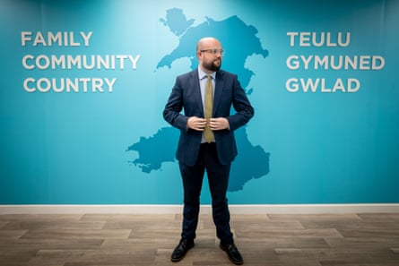 Powell stands in front of a turquoise background showing a map of Wales and the words ‘family, community, country’ in English and Welsh