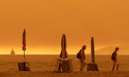 Two people trudge along a beach, with the sea behind them, and three folded beach umbrellas standing on the beach. The sky is a dark orange colour and everything in the picture is strongly tinted orange.