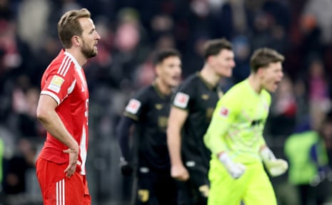 Bayern Munich Harry Kane looks dejected after the final whistle whilst Augsburg players celebrate in the background.