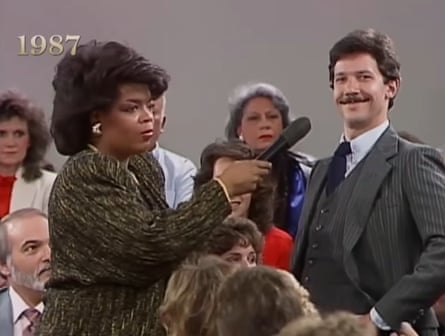 An angry Oprah holds a microphone to a smug-looking white man’s face as they stand in a crowd of all white people