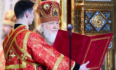 Patriarch Kirill in full regalia reads from a book held by an assistant