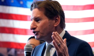 A man in a suit in front of a US flag speaks into a mic.
