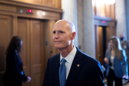Rick Scott, wearing a navy suit and blue tie, walks through a hall of the Capitol building.