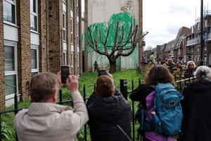 Crowds take pictures on their phones of a new Banksy artwork. It is a stencil of a person and spray-painted tree foliage on a wall behind a leafless tree