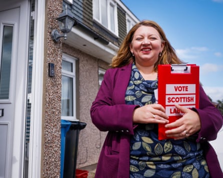Kirsty McNeill out campaigning in a residential street. She holds a red folder with the slogan Vote Scottish Labour. She is in her mid-40s with long reddish hair, and is smiling.
