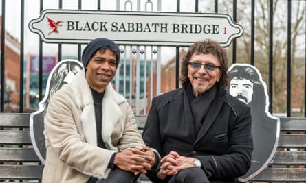 Carlos Acosta et Tommy Iommi assis à l'extérieur, les mains sur les genoux, sous un panneau indiquant « Black Sabbath Bridge » à Birmingham. 