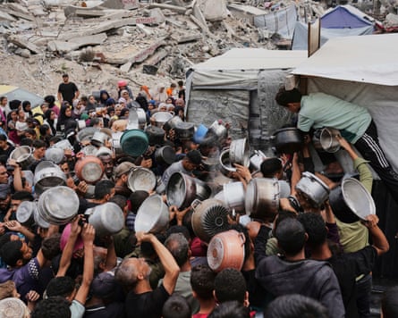 A crowd of people holding out empty containers towards a man distributing food