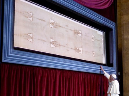 The pope reaching up to touch the wall-mounted case in which the Turin shroud is being displayed at its full 14-foot length
