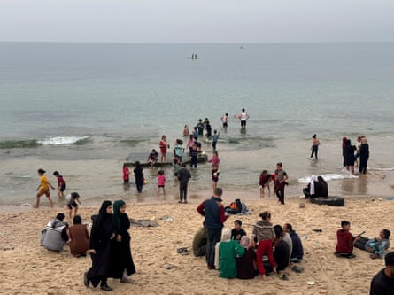Small groups of people sit on the sand while children and men wade into the water with their trousers rolled above their knees. A fishing boat is seen in the distance.
