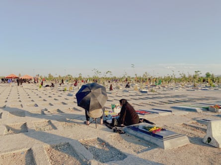 An unseen man on a chair with a black parasol and a woman dressed in black sit by a grave in Behesht-e Zahra cemetery in Iran.