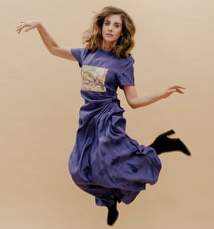 Alison Brie jumping, with arms and legs cocked, in a long, mauve T-shirt dress with full skirt gathered at the waist and heeled lack boots