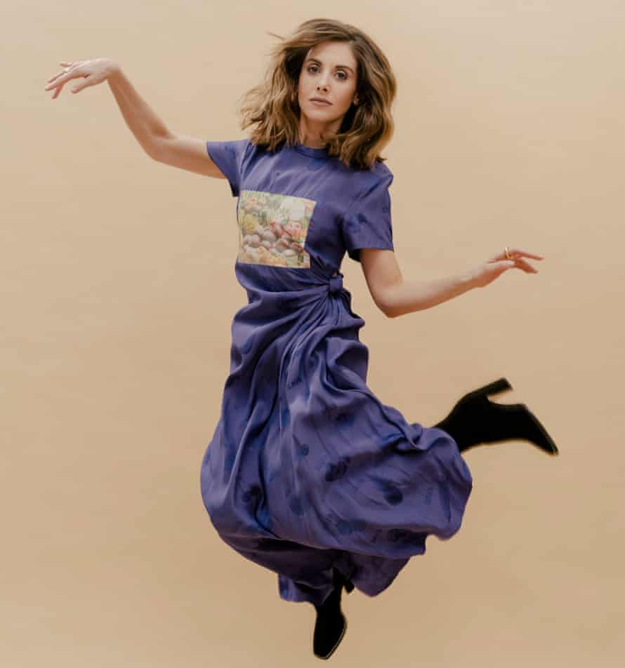 Alison Brie jumping, with arms and legs cocked, in a long, mauve T-shirt dress with full skirt gathered at the waist and heeled lack boots